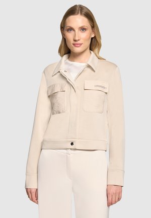 Frau mit blondem Haar trägt eine hellbeige Jacke mit zwei vorderen Klappentaschen und steht vor einem einfarbigen hellgrauen Hintergrund.