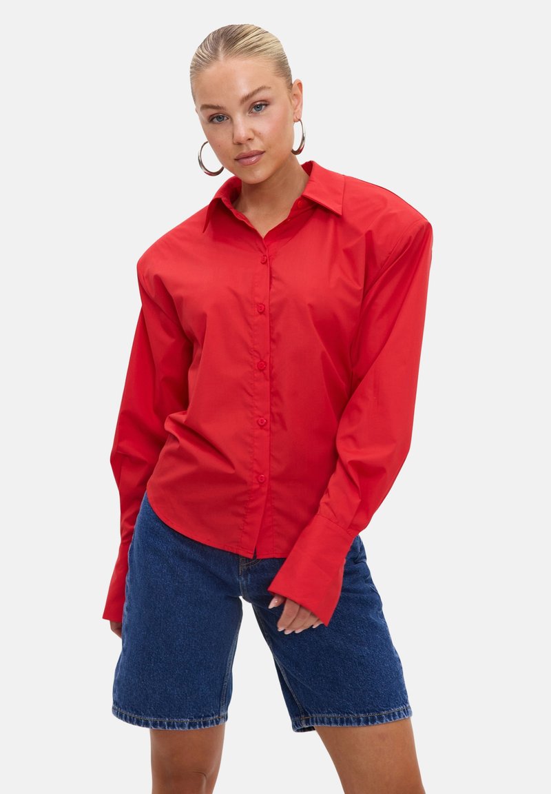 Femme aux cheveux plaqués en arrière porte une chemise rouge oversize boutonnée et un short en jean bleu longueur genoux, posant sur un fond uni.
