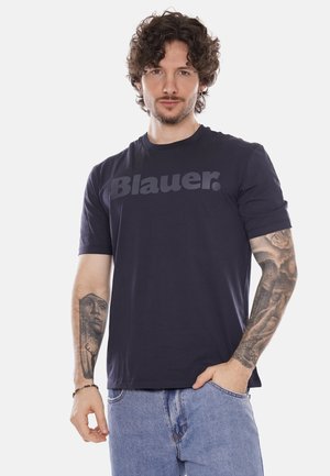 Uomo con capelli ricci che indossa una t-shirt Blauer blu navy e jeans azzurri, mostrando tatuaggi dettagliati su entrambi gli avambracci.