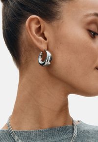 Silverfärgade, tjocka hoop-örhängen med en slät, reflekterande yta och en böjd design. Kan fästas med ett litet hoop-lås.