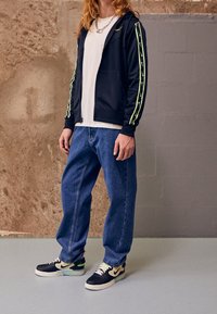 Černá mikina na zip s neonovými zelenými pruhy, pod ní bílý tričko, volné modré džínsy a černé/krémové tenisky se zelenými detaily.