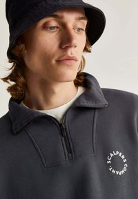 Navy sweatshirt met hoge kraag, half ritssluiting en wit cirkelvormig logo. Zachte textuur met een ontspannen pasvorm en eenvoudig ontwerp.