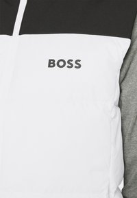 Juoda ir balta pūkinė liemenė su užtrauktuku, ant kurios priekyje juodai parašyta prekės ženklo pavadinimas "BOSS", kartu su pilku megztinio rankove.