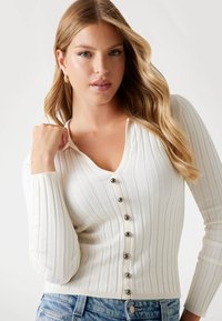Cardigan bianco a coste con scollatura a V, maniche lunghe e sei bottoni scuri sul davanti. Il materiale sembra morbido e aderente.