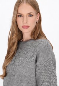 Grå strikket sweater med blomsterbroderi i et struktureret mønster. Har en rund halsudskæring og lange ærmer, der fremhæver håndværket.