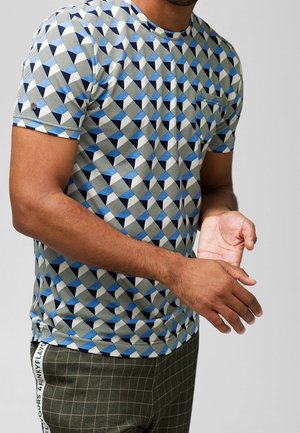Mann in einem kurzärmeligen T-Shirt mit blauem, schwarzem und weißem geometrischen Muster, kombiniert mit dunkelgrünen karierten Hosen mit seitlichem Textdetail.