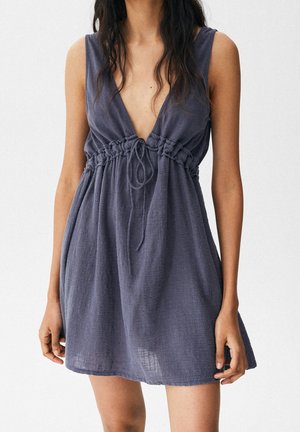 Vrouw draagt een mouwloze, wijde donkergrijze jurk met een diep V-hals, een geplooide taille en een strikdetail aan de voorkant.