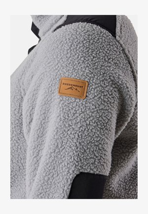 Grå fleecejakke med sorte skulderdetaljer. Har et lædermærke med teksten "SWEDEMOUNT" på venstre arm. Blød tekstur, afslappet design.