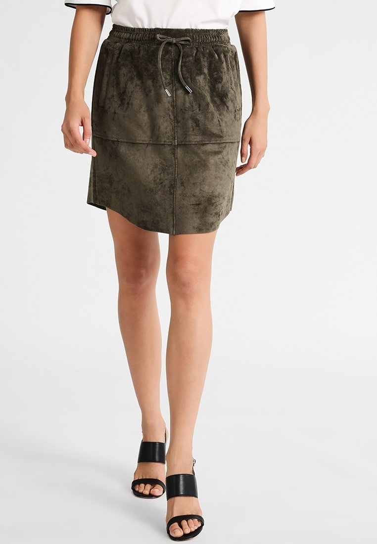 Femme portant une mini-jupe en daim vert olive avec une taille à cordon et des sandales à talons ouvertes noires, debout sur un fond blanc.