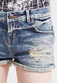 Szorty jeansowe w jasnoniebieskim kolorze z przetarciami, przeszyciami i podwiniętymi mankietami, z metalowym guzikiem i standardowymi kieszeniami.