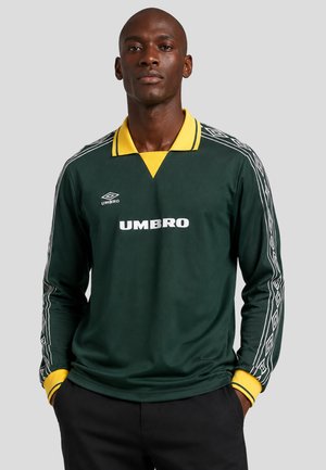 Uomo che indossa una maglia a maniche lunghe Umbro verde scuro con colletto e polsini gialli, in piedi con le mani nelle tasche davanti a uno sfondo grigio.