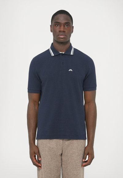 Navy polo shirt in cotton with a white striped collar. Features a small logo on the left chest. Worn with beige textured pants.