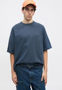 Mladý člověk v oranžové čepici, s přetáhnutým tmavě modrým tričkem a volnými modrými džínami na jednobarevném světlém pozadí.