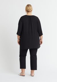 Schwarze, locker sitzende Tunika mit Dreiviertelärmeln, runder Halslinie und einem längeren Rückensaum, kombiniert mit schwarzen, engen Hosen und metallischen Sandalen.