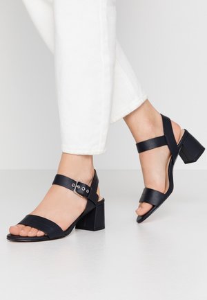 Fødder iført sorte læderhæle sandaler med ankelremme og sølv spænde, parret med hvide bukser mod en neutral baggrund.