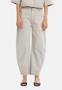 Jasnoszare dżinsowe spodnie z szerokimi nogawkami, kontrastowymi przeszyciami, kieszeniami z przodu i wysokim stanem, zestawione z czarnymi butami na otwartym palcu.