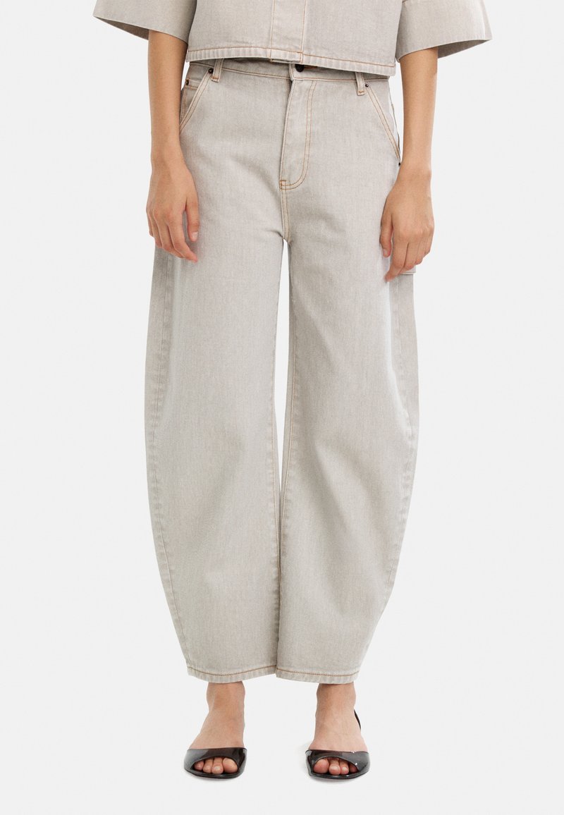 Jasnoszare dżinsowe spodnie z szerokimi nogawkami, kontrastowymi przeszyciami, kieszeniami z przodu i wysokim stanem, zestawione z czarnymi butami na otwartym palcu.