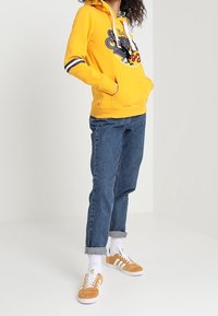 Sudadera amarilla con capucha y estampado gráfico, contrastada con rayas azul marino y blancas en las mangas. Combinada con jeans de denim azul remangados y zapatillas naranjas.