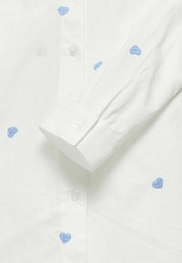 Camisa blanca con corazones bordados en azul; cuenta con cierre de botones y mangas con puños, hecha de un tejido ligero con una textura suave.