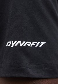 Κοντινή λήψη μαύρου υφάσματος με λευκό λογότυπο "DYNAFIT" τυπωμένο κοντά σε ραμμένη γαρνιτούρα.