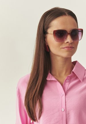Žena s dugom smeđom kosom, koja nosi velike sunčane naočale ljubičaste nijanse, crvene naušnice i ružičastu košulju na kopčanje, gleda blago udesno.