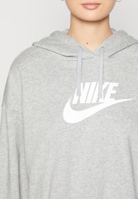 Sudadera gris hecha de un tejido suave con un bolsillo canguro, que presenta un gran logo de Nike en blanco en el frontal y cordones de color gris claro.