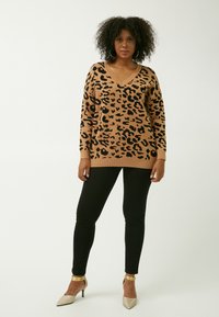 Maglione a maglia beige con motivo leopardato nero, scollo a V, maniche lunghe e orlo a coste, abbinato a leggings neri e tacchi con accenti metallici.