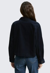 Persoon met lang bruin haar, gekleed in een donkerblauwe jas en een blauwe jeans, staande met de rug naar de camera tegen een effen lichtgrijze achtergrond.