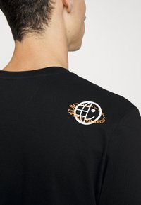 Czarna bawełniana koszulka z białym logo globusa oraz pomarańczowym i czarnym akcentem wzoru tygrysa na górze pleców. Widoczne krótkie rękawy.