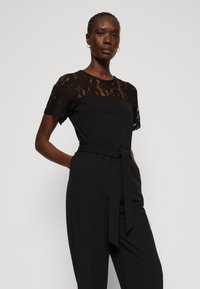 Combinaison noire avec un empiècement en dentelle et des manches courtes, dotée d'une taille cintrée par une ceinture et de poches latérales pour plus de détail et de fonctionnalité.