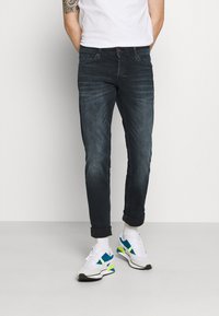 Calças de ganga escuras com um desgaste subtil, design de cinco bolsos e punhos enrolados, combinadas com sapatilhas brancas que apresentam detalhes em azul e verde.