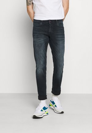 Calças de ganga escuras com um desgaste subtil, design de cinco bolsos e punhos enrolados, combinadas com sapatilhas brancas que apresentam detalhes em azul e verde.