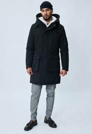Parka noir rembourrée avec capuche ajustable, deux poches avant, poche poitrine zippée et doublure blanche contrastante. Portée sur un pantalon gris.