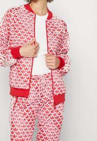 Röd och rosa bomberjacka med lövmönster, ribbade muddar och midjeband; matchas med byxor med liknande design och detaljer.