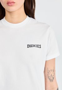 Bílý krátký rukáv tričko vyrobené z bavlny, s černým logem "DICKIES" na levé části hrudi. Hladká textura, kulatý výstřih.