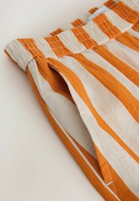 Nahaufnahme von orange-weiß gestreiften Stoffshorts mit elastischem Bund und sichtbarer Seitentaschenöffnung.