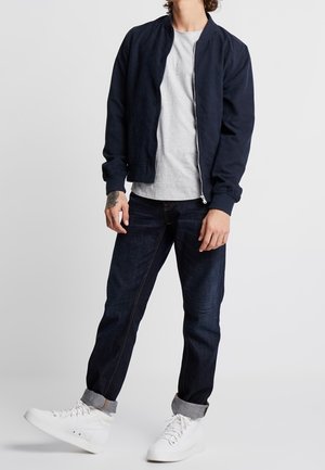 Chaqueta bomber de la Marina, camiseta gris, jeans de mezclilla oscura y zapatillas blancas. La chaqueta cuenta con un cierre de cremallera y puños de canalé.