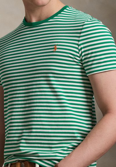 Roheline ja valge triibuline lühikeste varrukatega t-särk väikese oranži tikandiga logoga vasakul rinnal, kantud isiku poolt.