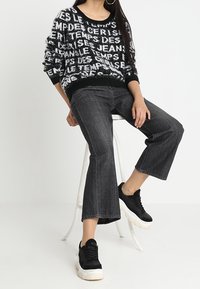 Svart tröja med vitt textmönster, i kombination med mörkgrå vida jeans och svarta plattformssneakers. Vit bakgrund och stol närvarande.