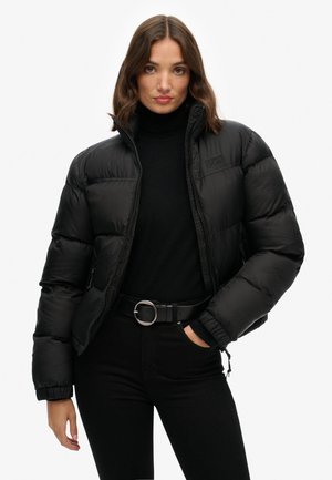 Schwarze Daunenjacke mit Reißverschluss, ausgestattet mit einem hohen Kragen und elastischen Bündchen. Getragen über einem schwarzen Rollkragenpullover und kombiniert mit schwarzen Hosen.