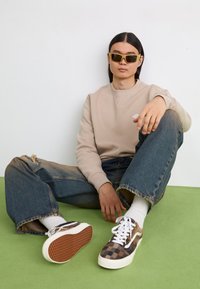 Beige hettegenser og lyseblå ripped jeans. Brune og hvite rutete sneakers. Person som sitter på grønn overflate, iført solbriller.