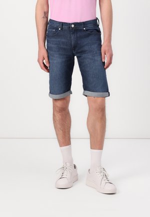 Shorts en denim bleu foncé, avec un ourlet roulé, des poches avant et une fermeture à bouton en métal. Associés à des baskets blanches et des chaussettes mi-mollet.