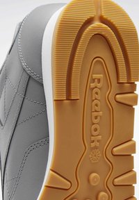 Harmaa tennari, jossa teksturoitu nahkapäällinen, valkoinen välikenkä ja kumipohja. Huomattava Reebokin brändäys pohjassa ja selkeä uritettu kuvio pohjassa.