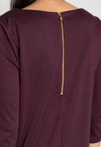 Robe bordeaux avec un col rond et une fermeture éclair dorée au dos. Le tissu est lisse et légèrement extensible, avec des manches trois-quarts.