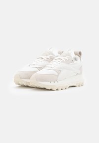 Pari valkoisia ja vaaleanbeigejä Reebok-tossuja, joissa on tekstuuripohjat ja nauhoitusdesign, tasaisella valkoisella taustalla.