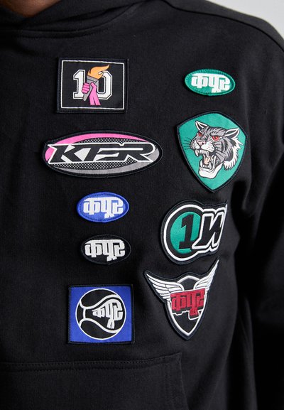 Μαύρη φόρμα με κουκούλα που διαθέτει διάφορα κεντημένα σχέδια: μια δάδα, τίγρη, λογότυπα σε ροζ, πράσινο, μπλε και λευκό, με διαφορετικά σχήματα και σχέδια.