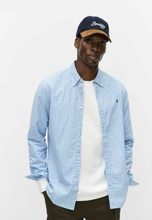 Man die een blauw geruit overhemd draagt over een witte trui, zwarte broek en een donkerblauw/beige baseballpet, staand tegen een effen achtergrond.