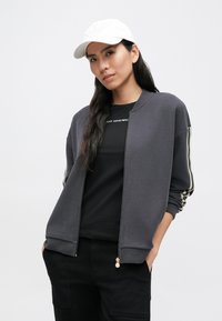 Szara kurtka bomber z zamkiem błyskawicznym i mankietami, z paskowanymi akcentami na rękawach. W zestawie z czarną koszulką i białą czapką.