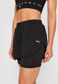 Bermudas deportivas negras con un diseño en capas, que presentan una cintura elástica y aberturas laterales, combinadas con pantalones cortos de compresión negros debajo.