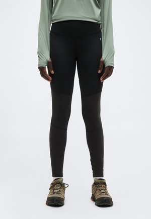 Άτομο που φοράει μαύρα κολάν ψηλής μέσης, πράσινο χρώμα φασκόμηλου μακρυμάνικο μπλουζάκι και μπεζ παπούτσια πεζοπορίας με μαύρες σόλες, στέκεται σε λευκό φόντο.